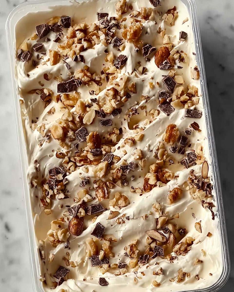 A rectangular plastic container filled with a creamy white base, the top spread unevenly with soft swirls and small peaks. Scattered generously across the surface are chopped nuts and bits of dark chocolate, providing a mix of light brown, beige, and deep brown colors with rough, uneven textures. The container is placed on a white marbled surface, visible mostly at the edges. The entire image is bright and clear, focusing closely on the creamy layered dessert. photo taken with an iphone --ar 4:5 --v 7