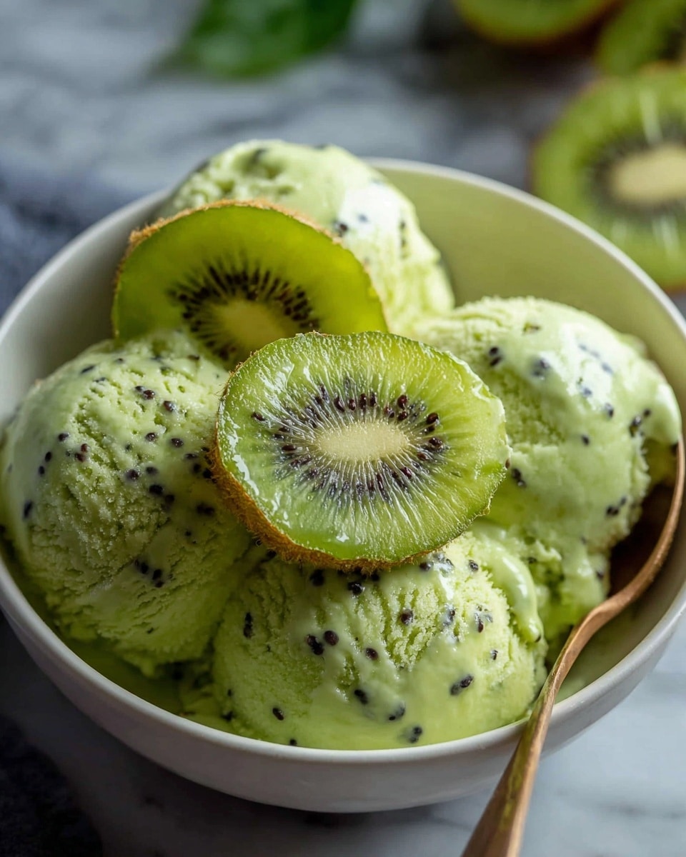 A close-up of a white bowl filled with three bright green ice cream scoops mixed with black kiwi seeds on the bottom layer. On top and around the scoops, there are several round slices of fresh green kiwi fruit with tiny black seeds and a lighter green center. A bronze spoon is placed inside the bowl on the right side. The bowl sits on a white marbled surface with some blurred green leaves in the background. Photo taken with an iphone --ar 4:5 --v 7