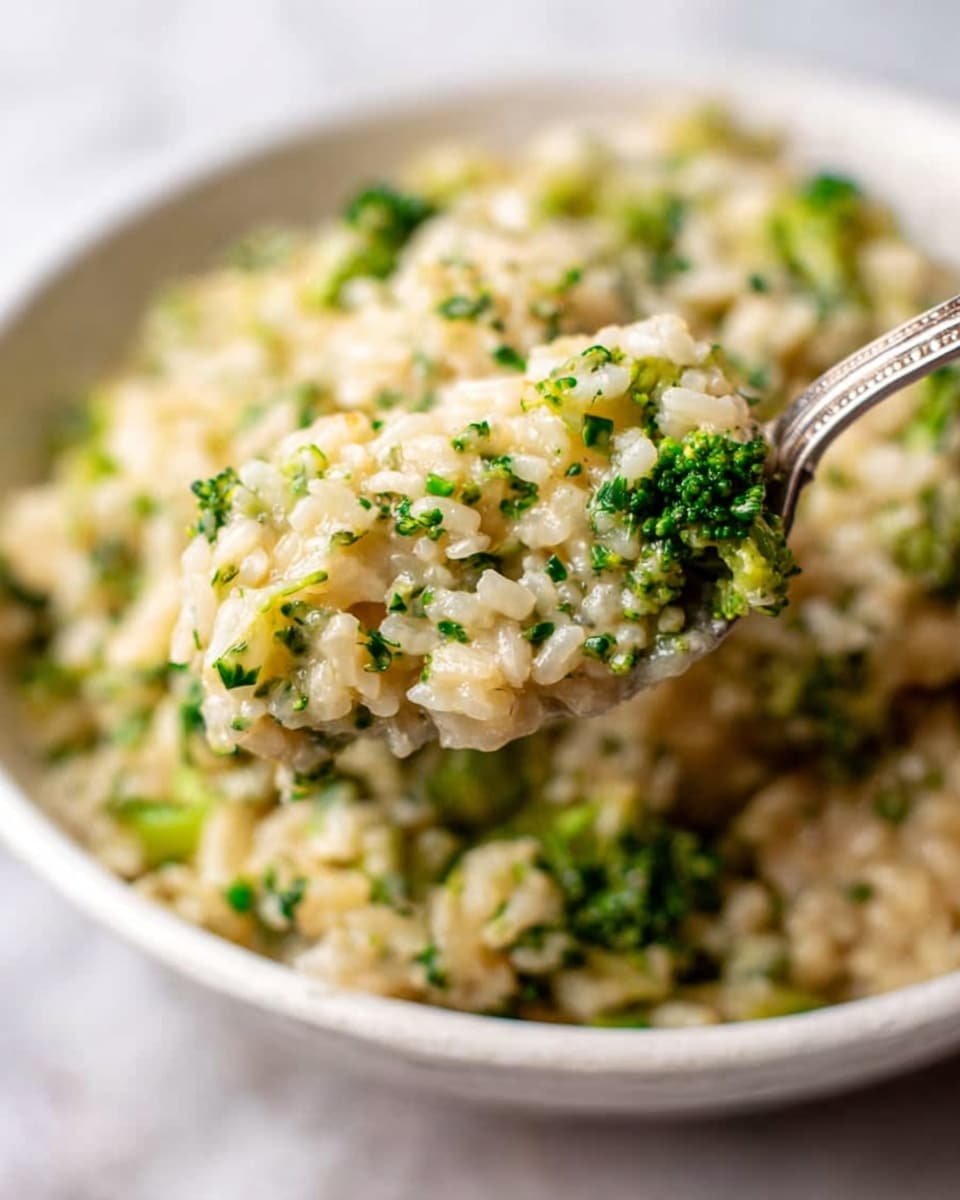 The image shows a close-up of a white bowl filled with creamy rice mixed with small green broccoli pieces, giving a textured look with soft, cooked grains and tiny florets spread evenly. A silver metal fork holds a scoop of the mix, with the grains and broccoli clearly visible, highlighting a smooth yet slightly chunky texture. The bowl sits on a white marbled surface that adds a clean and bright background to the warm, inviting dish. photo taken with an iphone --ar 4:5 --v 7