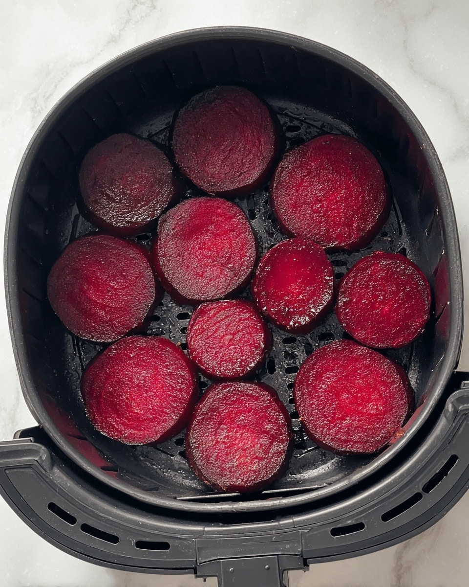 The image shows a black air fryer basket with ten round slices of deep red beetroot placed in one layer inside, each slice showing a smooth, moist surface with a slightly shiny texture. The basket has a perforated metal base that is visible between the beetroot slices. The background is a white marbled texture. photo taken with an iphone --ar 4:5 --v 7
