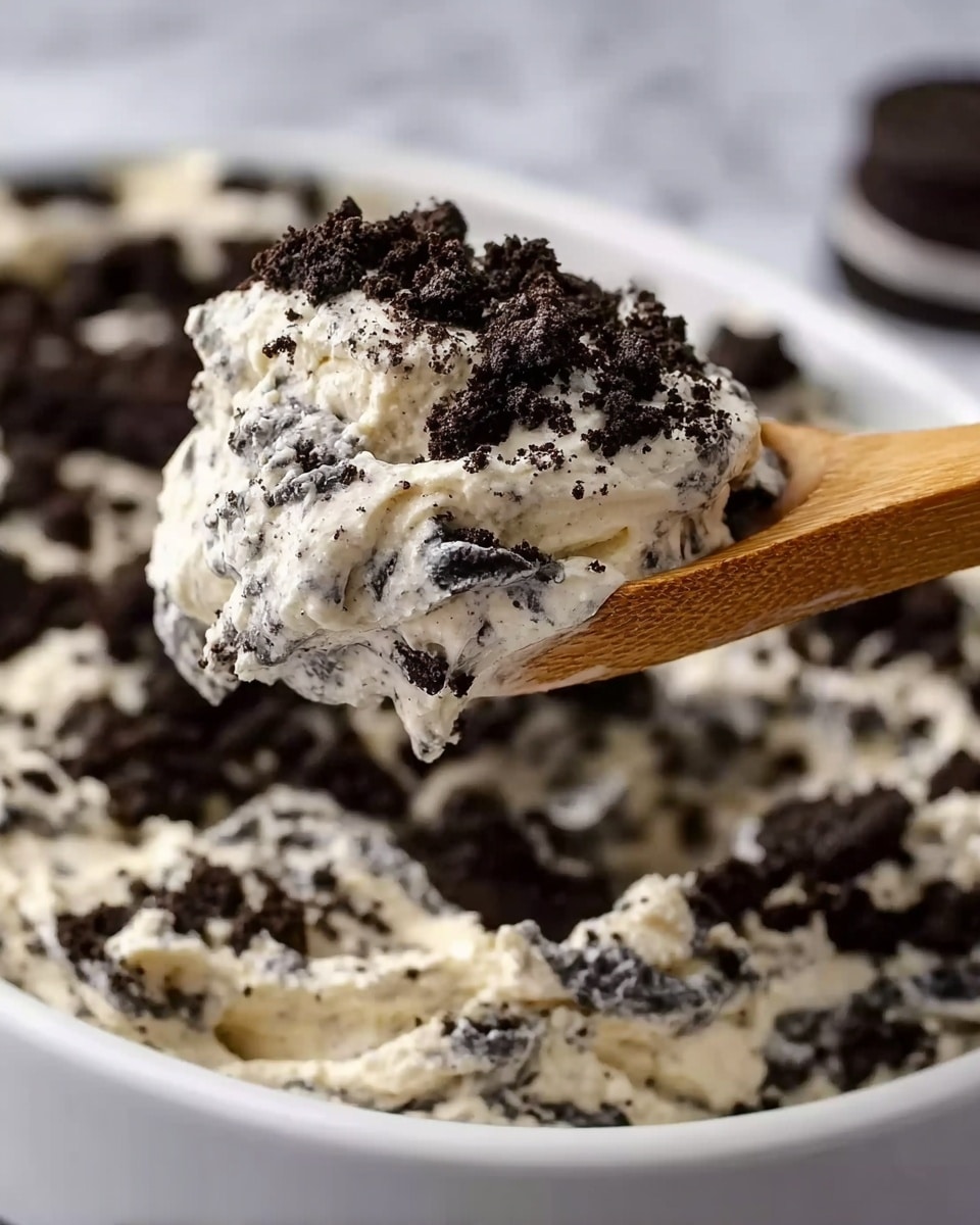 A close-up view shows a wooden spoon holding a scoop of creamy dessert with visible black cookie pieces mixed evenly throughout. The background shows a large white bowl filled with the same creamy mixture topped with a generous layer of crumbled black cookies. The texture of the cream looks smooth and soft with dark cookie chunks creating a rough contrast. The setting has a white marbled surface in the background. photo taken with an iphone --ar 4:5 --v 7