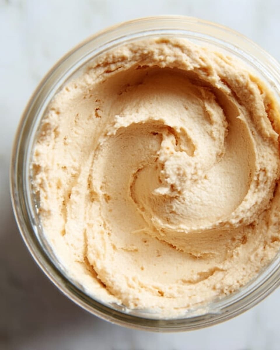 A close-up view of light beige creamy spread with smooth, fluffy texture shown inside a transparent glass jar, with gentle swirls on the surface and a small scoop mark near the center, all placed on a white marbled surface photo taken with an iphone --ar 4:5 --v 7