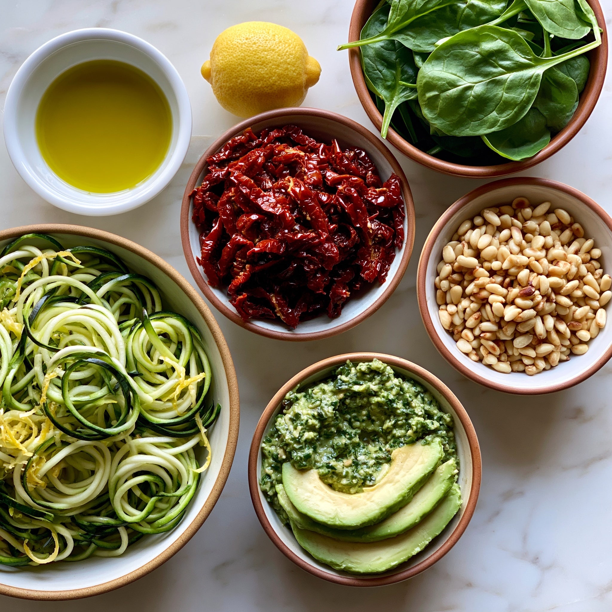 Two white round plates each hold a serving of green pasta twisted into small nests. The pasta is coated with a creamy green sauce and mixed with wilted dark green spinach leaves and pieces of red sun-dried tomatoes. Each plate is garnished with a wedge of yellow lemon and a sprinkle of light brown pine nuts. A silver fork rests on the bottom plate’s edge. Nearby there is a halved avocado with bright green flesh, two clear glasses filled with a pale yellow drink, a small white bowl of pine nuts, and fresh green basil leaves on a white marbled surface. A dark teal cloth is partly visible on the left side. photo taken with an iphone --ar 4:5 --v 7