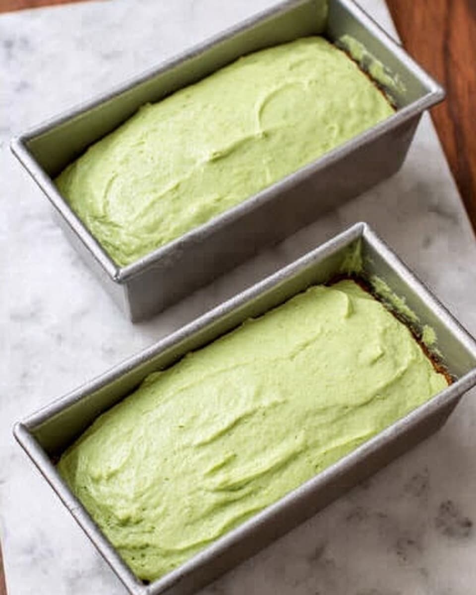 The image shows two silver loaf pans filled with light green batter that is smooth and softly textured. The batter evenly fills each pan with no additional layers visible. The pans are placed on a white marbled surface that adds a clean, subtle pattern to the background. The light green color looks creamy and fresh, suggesting a dessert or bread with matcha or pistachio flavor. Photo taken with an iphone --ar 4:5 --v 7