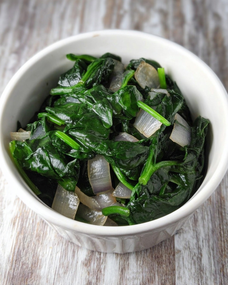 A white bowl filled with a mix of cooked spinach leaves and diced translucent gray onions. The spinach leaves are dark green and glossy, layered evenly with the soft, slightly shiny onion pieces throughout. The bowl sits on a white marbled surface, showing a simple and clean presentation. Photo taken with an iphone --ar 4:5 --v 7