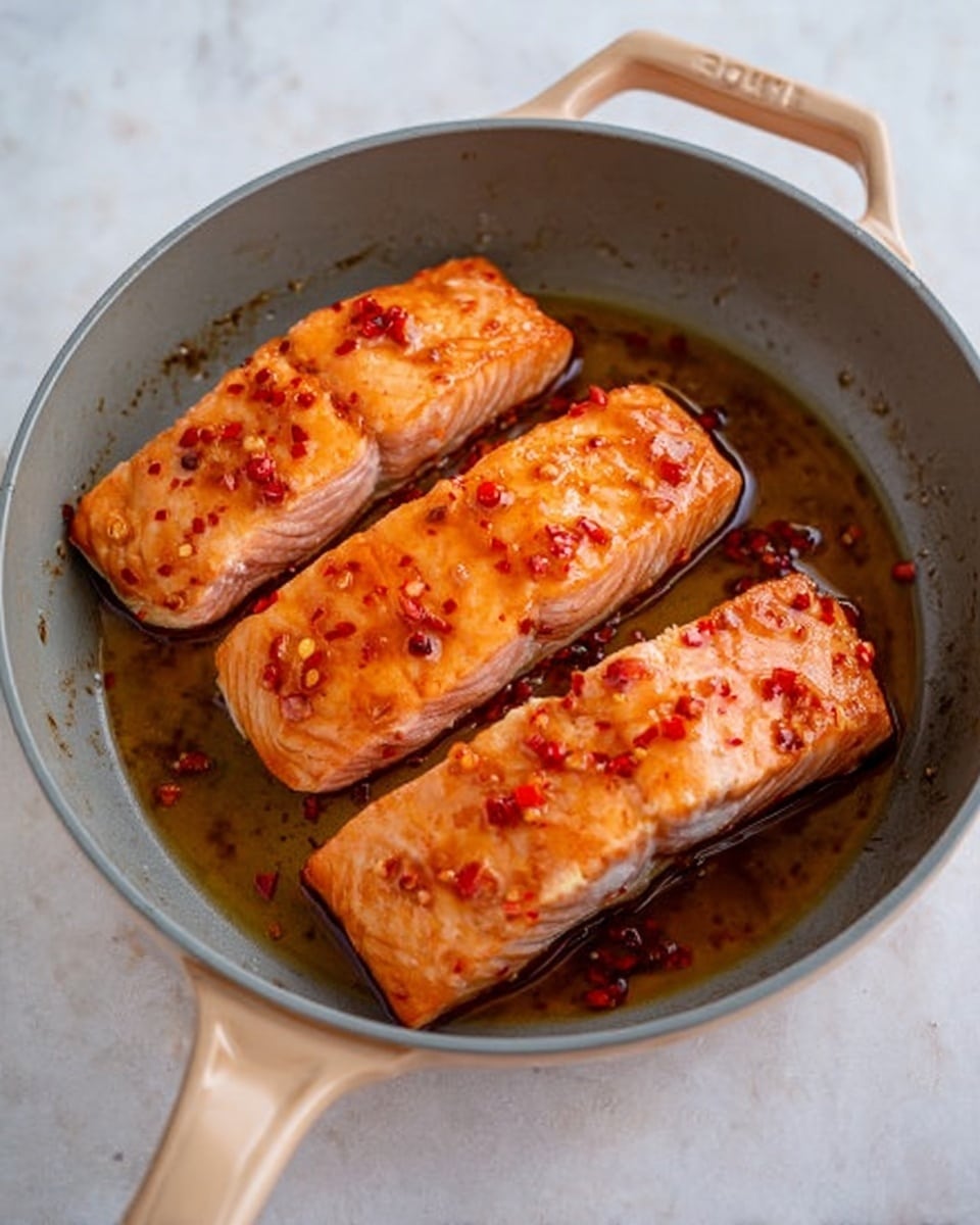 The image shows a gray pan with light brown handles on a white marbled surface. Inside the pan, there are three pieces of cooked salmon with a shiny, slightly orange glaze that has small red chili flakes on top. The salmon pieces have light pink sides and a crispy orange-brown top layer. There is a small amount of oil around the salmon, making it look juicy and hot. Photo taken with an iphone --ar 4:5 --v 7