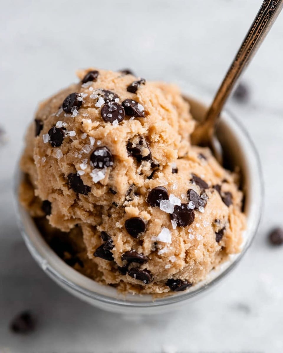 A small white bowl filled with two scoops of light brown cookie dough mixed with many small dark chocolate chips, topped with large flakes of sea salt. A thin metal spoon is stuck into the cookie dough, with its handle going out of the frame. The bowl sits on a white marbled surface with a few chocolate chips scattered around. The lighting is soft and natural, highlighting the texture of the dough and chocolate chips. photo taken with an iphone --ar 4:5 --v 7