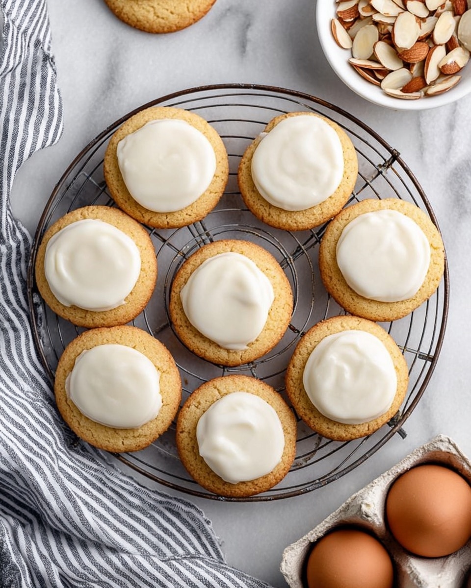 Eight soft round cookies with smooth white icing in the center are placed on a round wire cooling rack. One more cookie without icing sits partially off the rack near a white egg carton holding three brown eggs at the bottom right. A small white bowl filled with sliced almonds is near the top right side. Part of a striped cloth is visible on the bottom left side. The background is a white marbled texture. photo taken with an iphone --ar 4:5 --v 7