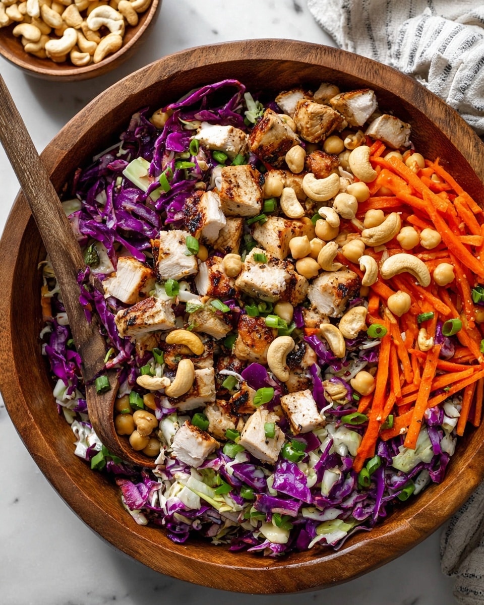 The image shows a large wooden bowl filled with a colorful salad. The salad has several layers, starting with chopped purple cabbage that adds a deep purple color and slightly rough texture. On top, there are bright orange carrot sticks spread evenly. Scattered around are small, round, beige chickpeas, mixed with chunks of grilled white chicken pieces that have golden-brown marks and a slightly crispy outside. Green chopped spring onions are sprinkled all over, adding a fresh look. Large beige cashew nuts are spread through the salad for a crunchy texture. A wooden spoon rests inside the bowl on the left side, partly buried under the salad. In the background, there is a small wooden bowl with more cashews and a white marbled surface under everything. The photo taken with an iphone --ar 4:5 --v 7