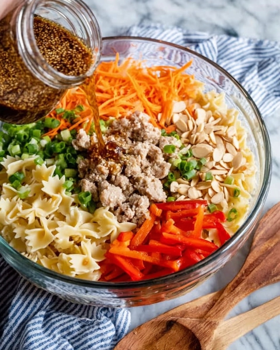 A large clear glass bowl holds several layers of ingredients arranged in separate sections: pale yellow cooked pasta bows at the bottom left; bright orange shredded carrots at the top left; small chunks of light brown cooked meat at the top center; finely chopped green onions next to the meat towards the middle; thinly sliced red bell peppers at the bottom right; and a cluster of beige sliced almonds at the top right. A woman's hand pours a brown sauce filled with sesame seeds from a glass jar into the center of the bowl. The bowl sits on a white marbled surface, and a blue and white striped cloth and wooden spoon lie in front. Photo taken with an iphone --ar 4:5 --v 7
