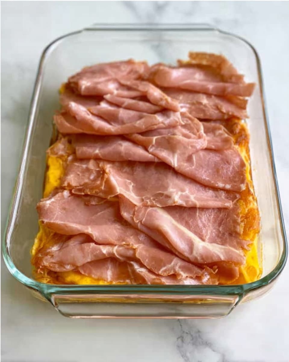 A clear rectangular glass dish shows two layers: the bottom layer is a bright yellow, likely cheese or sauce, spread evenly across the base. The top layer consists of several slices of soft, pinkish-brown meat arranged with slight folds and overlapping edges, filling the dish entirely. The dish rests on a white marbled surface. photo taken with an iphone --ar 4:5 --v 7