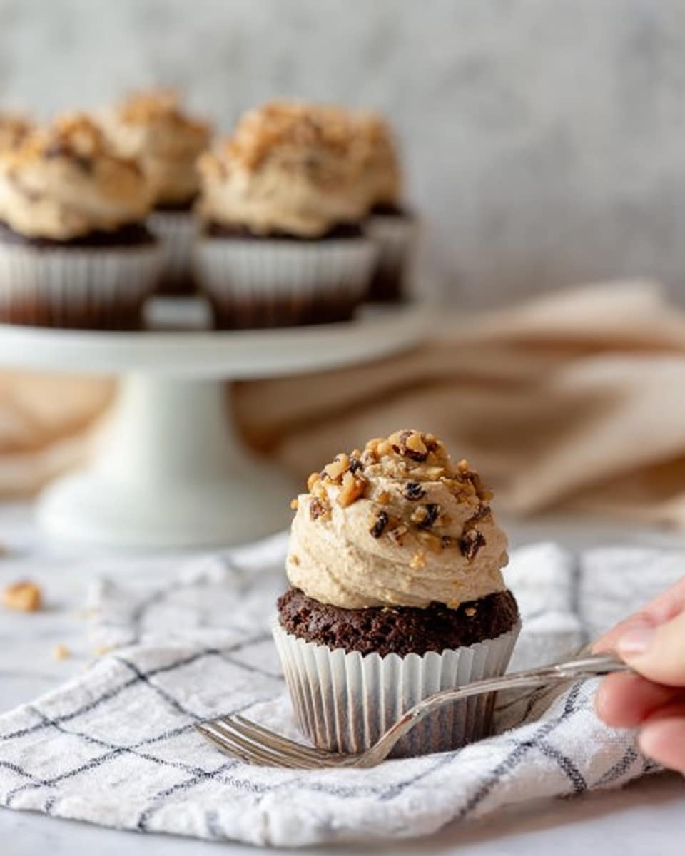 A white paper cup holds a dark brown chocolate cupcake topped with a large scoop of light beige cookie dough frosting that has small dark chocolate bits and chopped walnuts on top. This cupcake sits on a white cloth with a black grid pattern, next to a silver fork with a woman’s hand gently holding it. In the background, there is a white marble stand with more cupcakes like the one in front, all topped with the same frosting and walnuts. Behind the stand, a soft beige cloth is casually draped on a white marbled surface. Photo taken with an iphone --ar 4:5 --v 7