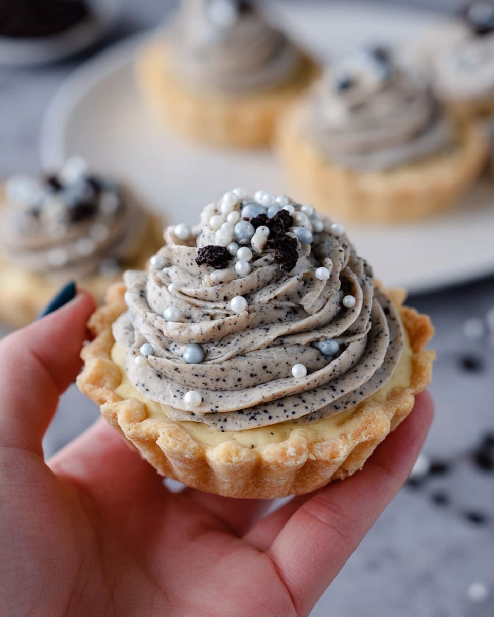A single dessert sits on a dark plate with a white marbled surface underneath, featuring two layers: the bottom layer is a pale yellow, rough-textured base that looks soft and slightly crumbly; on top is a swirled mound of light grey cream with small dark specks, shaped like a soft peak. Scattered across the cream are small, smooth, round beads in white, light blue, and black colors, giving a decorative touch. More similar desserts can be seen softly blurred in the background on stacked white plates. Photo taken with an iphone --ar 4:5 --v 7