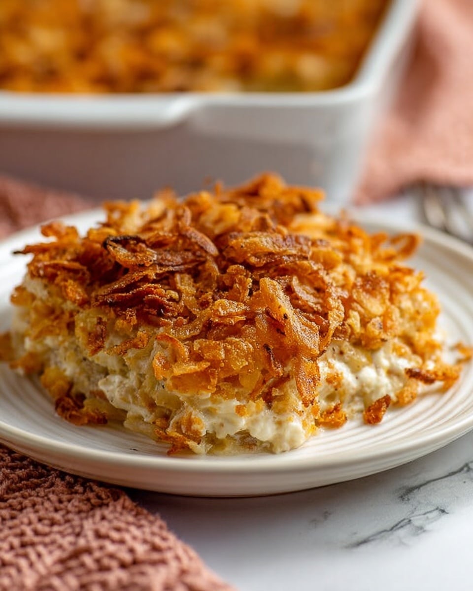 A large serving of a casserole sits on a white plate with a white marbled surface underneath. The casserole looks like it has three layers: the bottom layer is creamy white with small pieces mixed inside, the middle layer is thick and mixed with melted cheese, and the top layer is golden brown with crunchy, crispy fried onions covering the whole surface. The casserole edges show a mix of creamy and crunchy textures, and the colors range from white to light brown and golden orange. In the background, the same casserole can be seen in a white rectangular dish. Nearby, a woman's hand wearing a pink knitted cloth is visible at the edge of the image. photo taken with an iphone --ar 4:5 --v 7