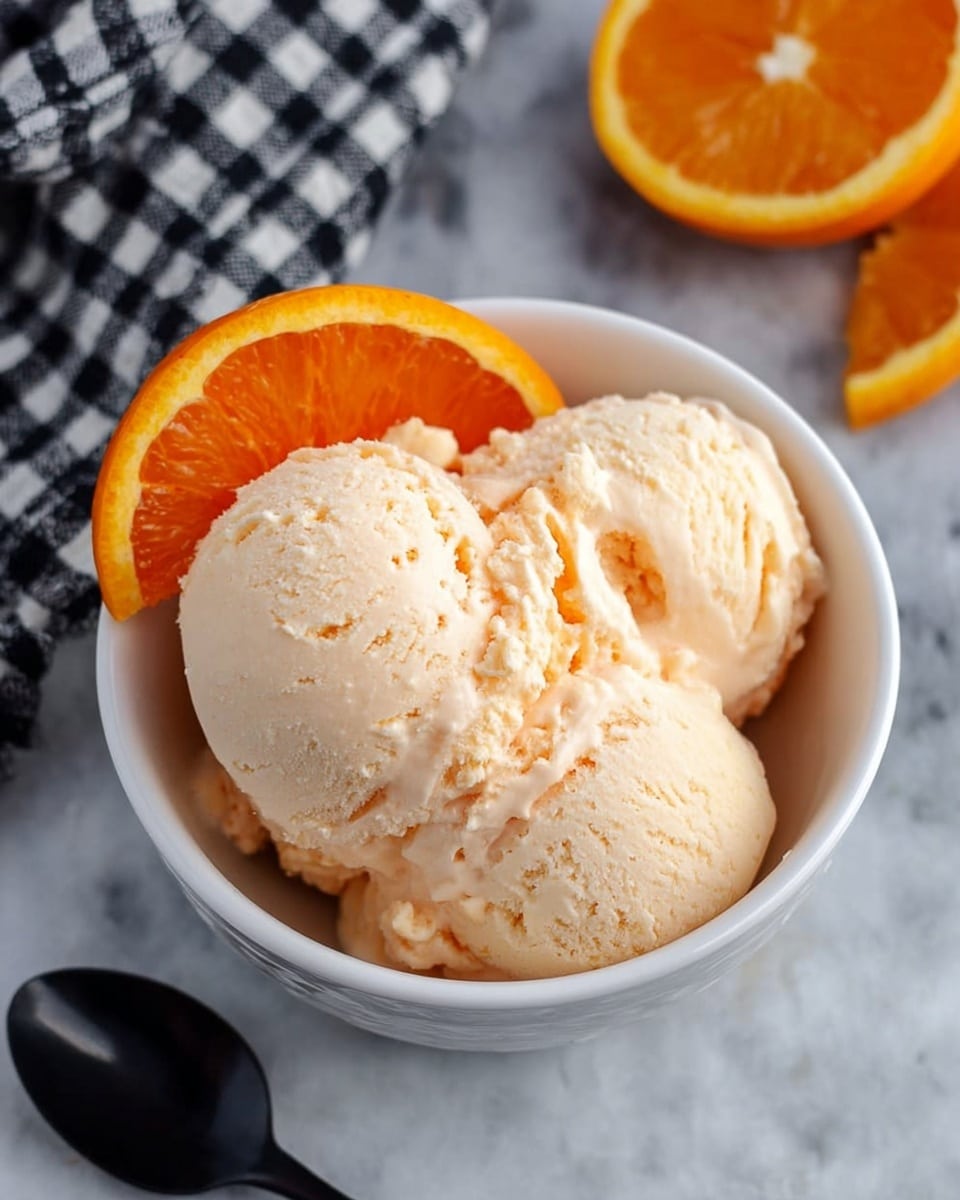 A white bowl filled with three scoops of light peach-colored ice cream, topped with a bright orange slice resting on the edge. The texture of the ice cream looks creamy and slightly rough. The bowl sits on a white marbled surface next to a black spoon, with a black and white checkered cloth in the top left corner and a half orange slice in the top right corner. photo taken with an iphone --ar 4:5 --v 7