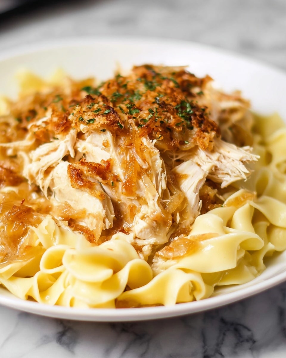 The dish shows a pile of wide egg noodles at the bottom with a light yellow color and smooth texture, topped by layers of cooked shredded chicken that is white with a slightly fibrous texture. On top of the chicken, there are caramelized onions with a golden brown shade and soft texture covering parts of the dish. A crispy layer of golden brown baked topping with some herbs sprinkled is laid on the very top, adding crunch and color contrast. The plate is white and the background is a white marbled texture. photo taken with an iphone --ar 4:5 --v 7