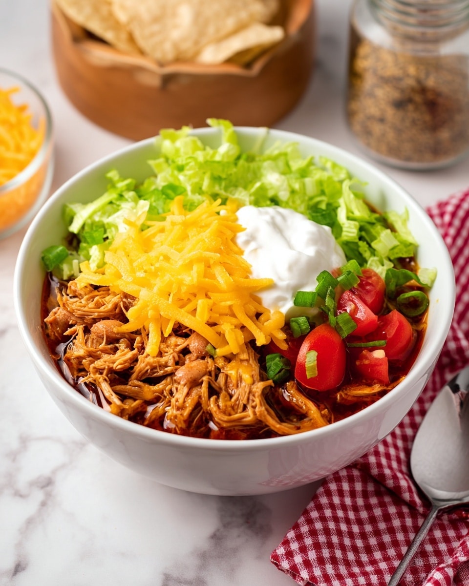 A white bowl filled with shredded cooked chicken mixed with light brown pinto beans in broth as the bottom layer. On top, shredded yellow cheddar cheese and thinly sliced pale green lettuce are scattered. A dollop of white sour cream sits in the center, surrounded by small red diced tomatoes and chopped green onions. The bowl sits on a white marbled surface with a red and white checkered cloth and a shiny silver spoon next to it. In the background, there is a wooden bowl holding round light beige tortilla chips, a glass jar of spices, and a block of yellow cheddar cheese with a grater nearby. Photo taken with an iphone --ar 4:5 --v 7