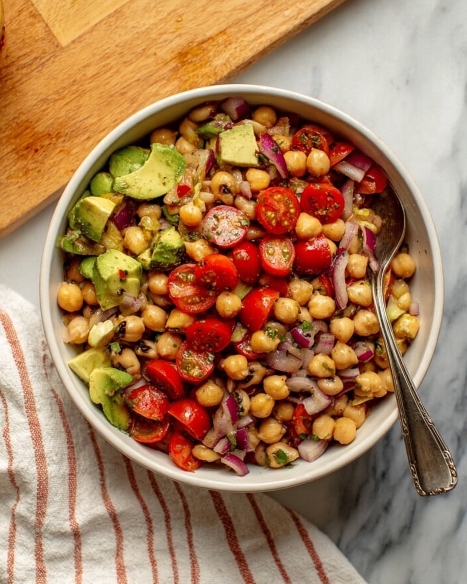 A white bowl filled with a colorful bean salad, showing three main layers: a base of small beige chickpeas and black-eyed peas mixed with chopped red onion and red bell pepper, a middle layer of chunks of green avocado, and a top layer featuring halved, bright red cherry tomatoes. The salad is mixed with small bits of green herbs and has a shiny, slightly wet texture, suggesting a light dressing. A silver spoon is placed inside the bowl on the right side. The bowl sits on a white marbled surface with a wooden board in the top left and a white cloth with light red stripes on the right side near the bowl. Photo taken with an iphone --ar 4:5 --v 7
